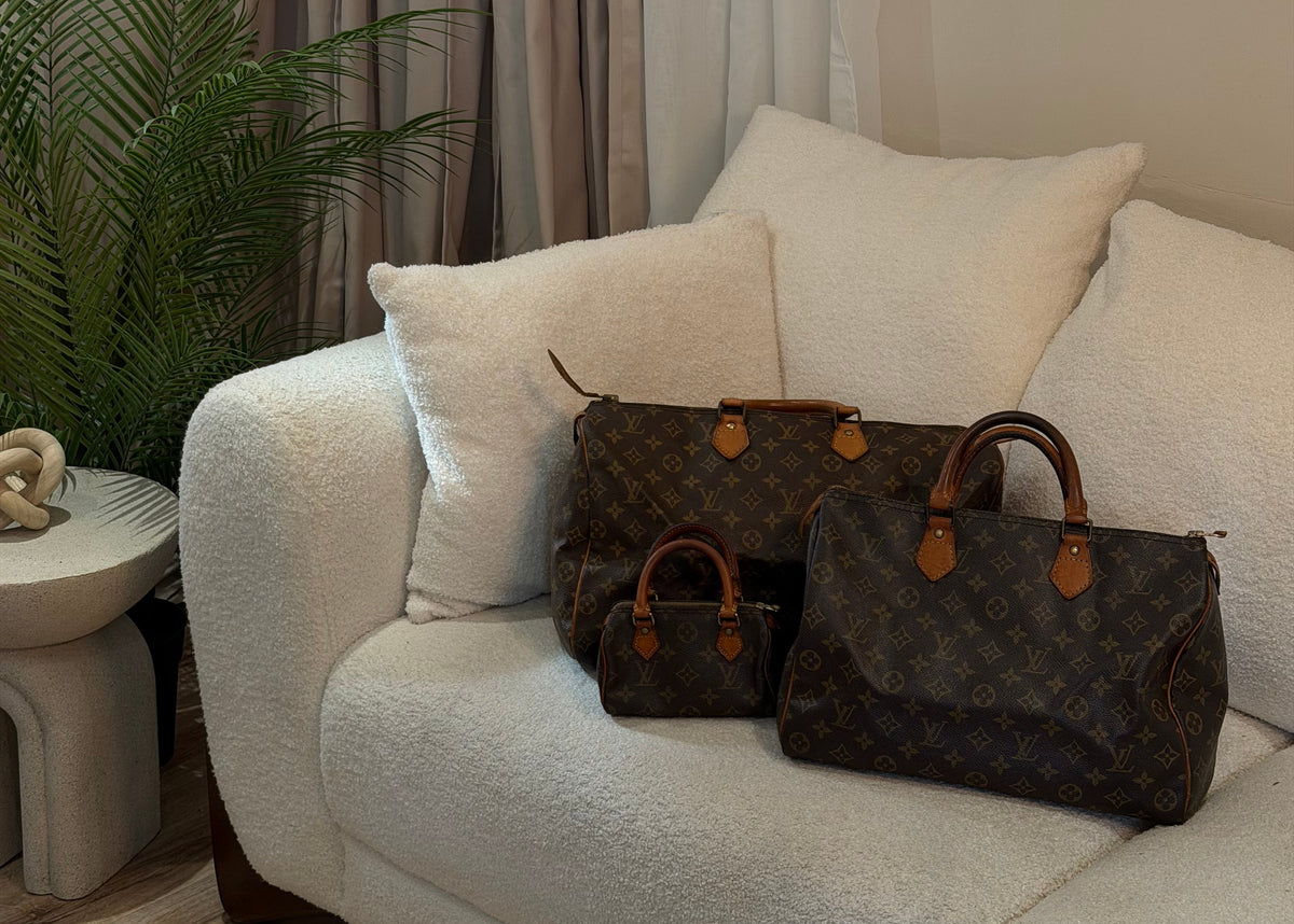 Three Louis Vuitton monogram handbags in different sizes arranged on a cream boucle sofa in a styled living room setting.